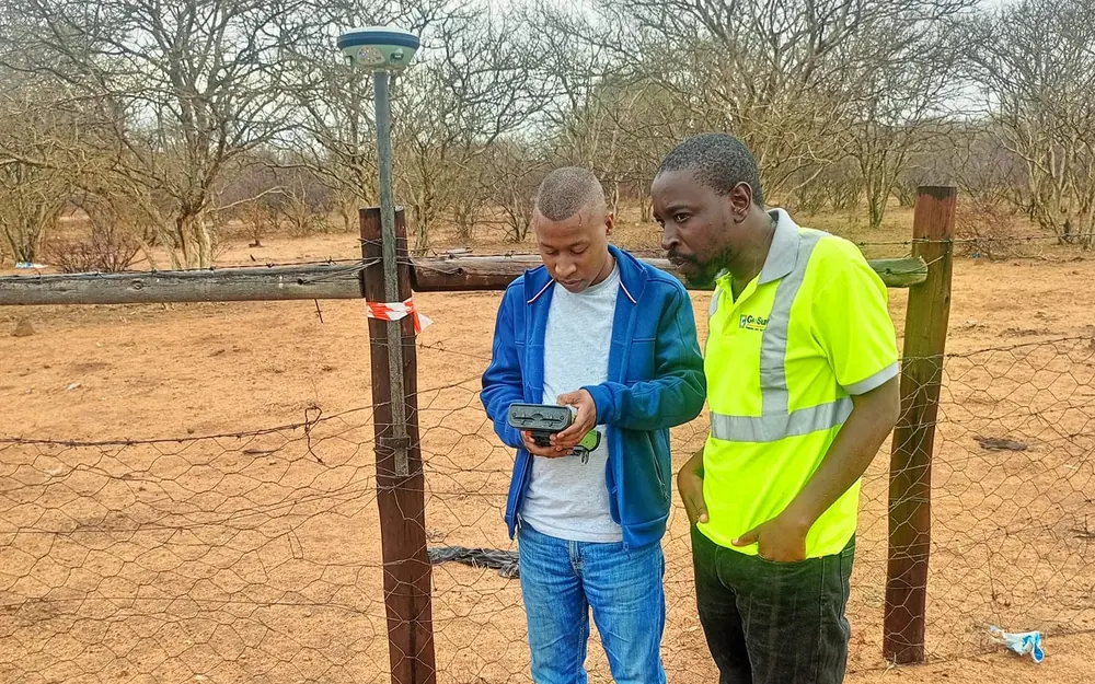 Two surveyors checking GNSS survey data on a handheld controller beside a fence line